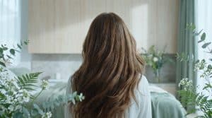 Vue de dos d'une personne aux longs cheveux bruns ondulés et brillants, entourée de feuillage vert et de fleurs blanches dans une pièce lumineuse.